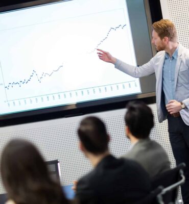 a man standing in front of a screen pointing at a graph